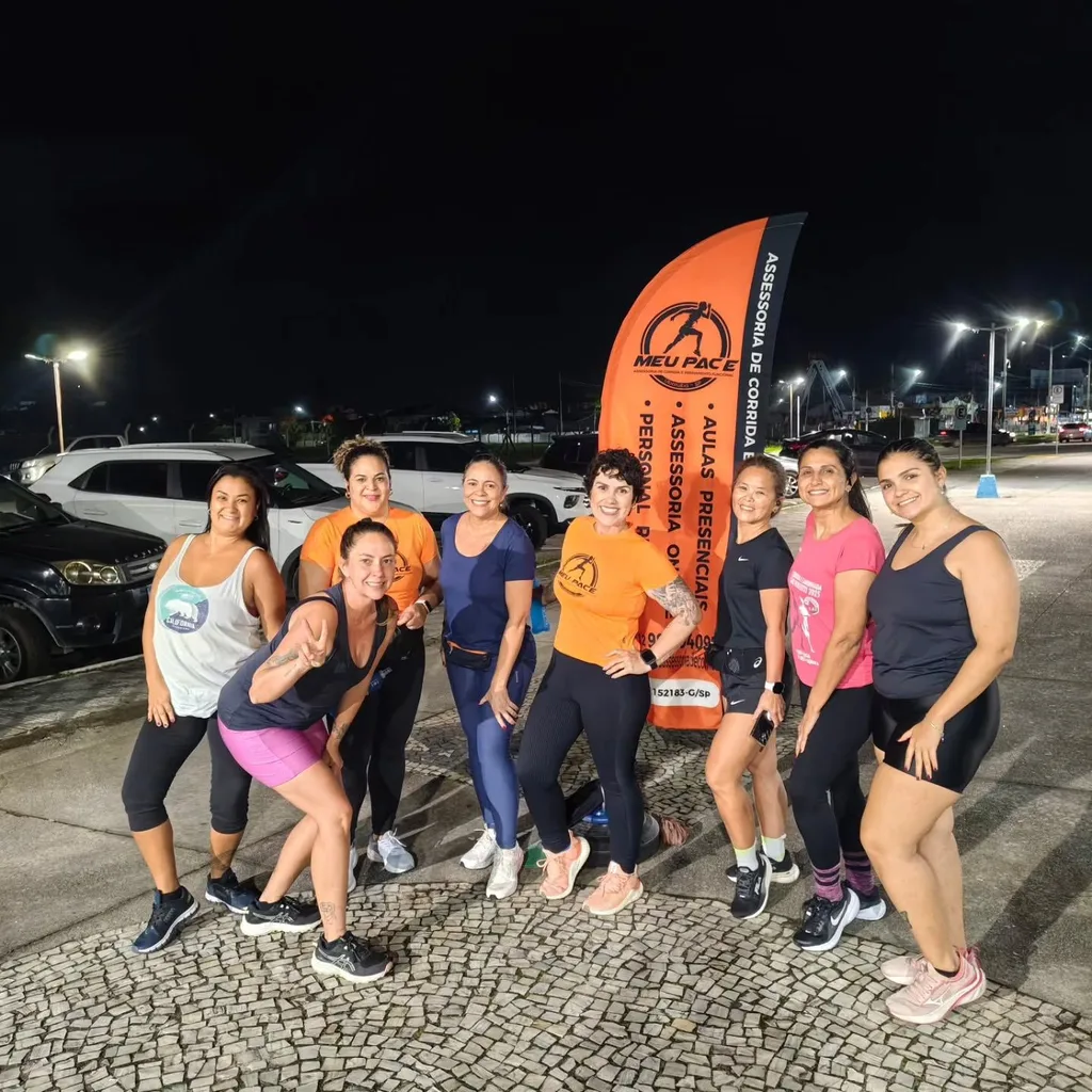 Grupo de alunas da Meu Pace à noite, na orla de Ubatuba, com a bandeira laranja da assessoria